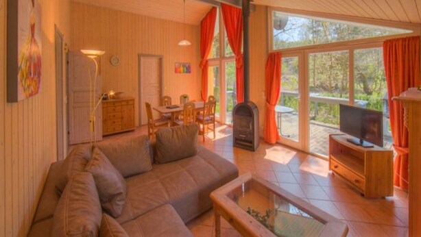A cozy living room with a sofa, a coffee table, and a fireplace. There is a dining area with a table and chairs, and a television on a wooden stand. The room has large windows with curtains and a view of the outdoors. - Ferienhaus mit Sauna im Ferienpark Mirow (Mirow)