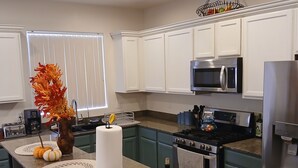 Uma cozinha moderna com armários brancos, uma ilha central com banquetas, micro-ondas, frigorífico e fogão.