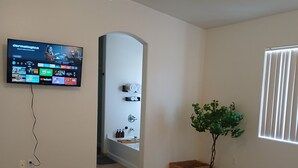 Quarto de hotel com cama, televisão na parede, banheiro com chuveiro e planta em vaso.