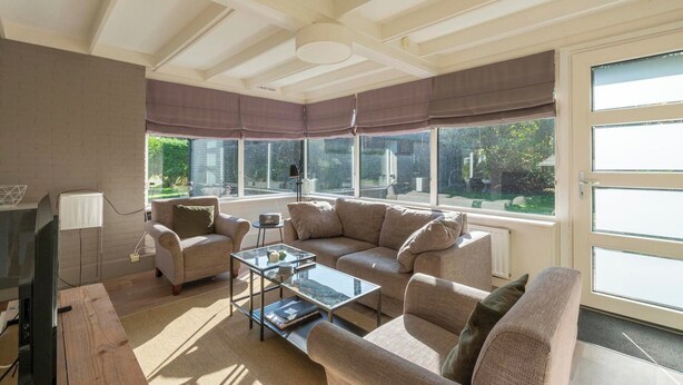 A living room with a sectional sofa, a glass coffee table, and a television. There are large windows with blinds, a door, and a radiator. - Bungalow in Sint Maartenszee Near Beach (Sint Maartensvlotbrug)