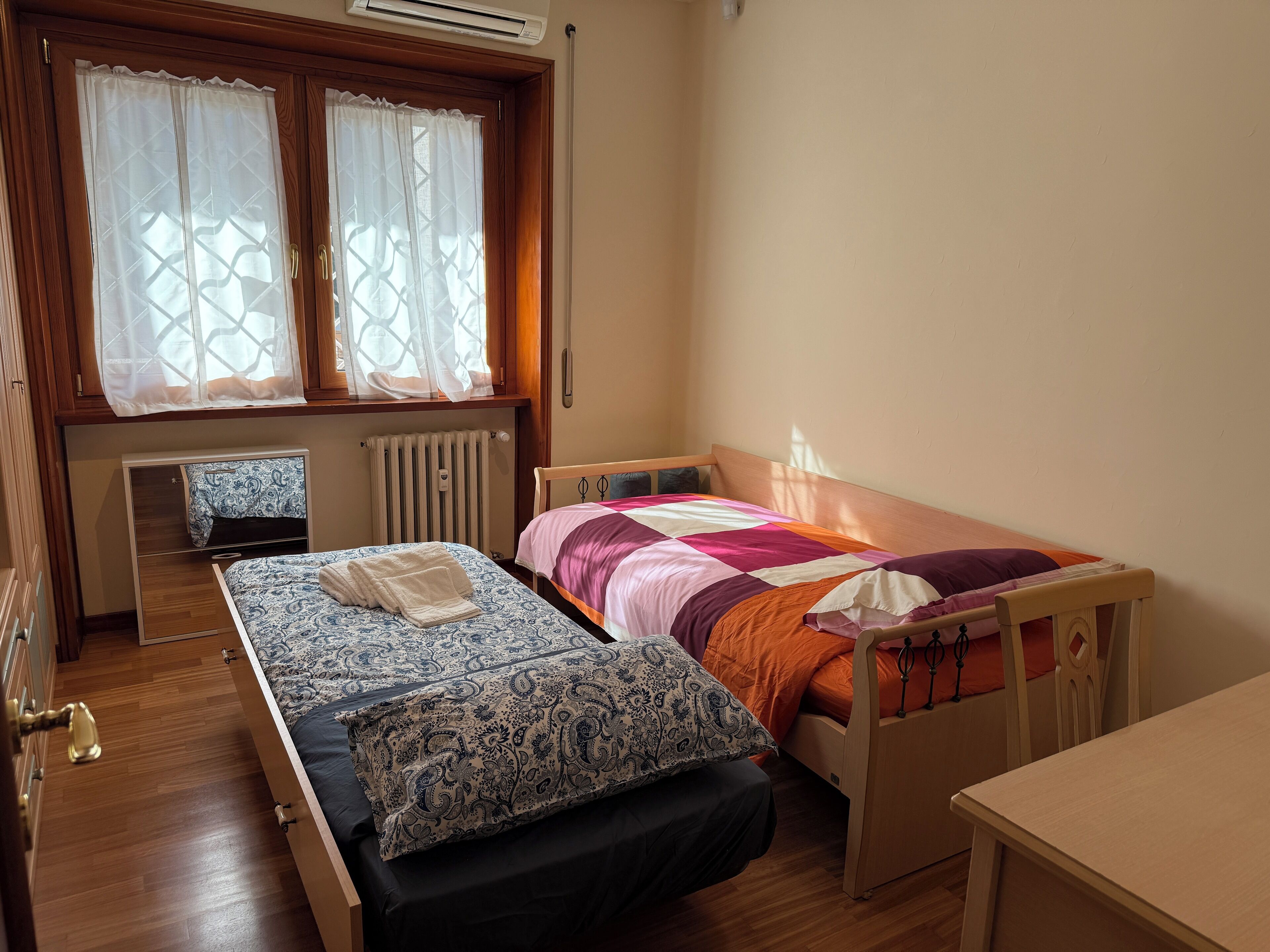 A small, neatly arranged bedroom with a single bed, a wooden nightstand, a small table, and a window with curtains.