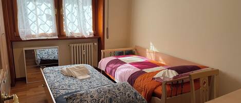 A small, neatly arranged bedroom with a single bed, a wooden nightstand, a small table, and a window with curtains.