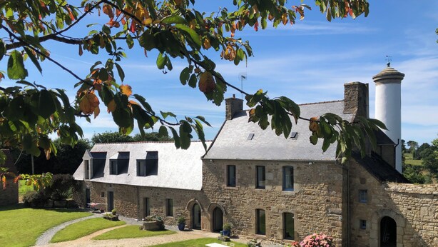 A stone building with a white roof, a chimney, and a white tower. There is a garden with flowers and a pathway leading to the building. - Sous Le Phare (Louannec)