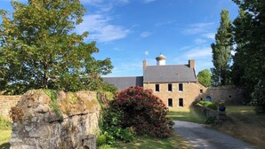 A stone building with a central chimney, surrounded by greenery and a paved path. - Sous Le Phare (Louannec)