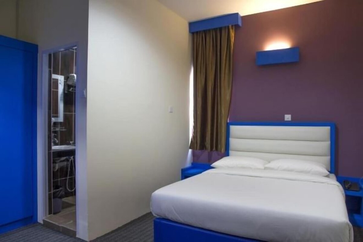 A hotel room with a bed, a nightstand, a wall-mounted light, and a door leading to a bathroom.