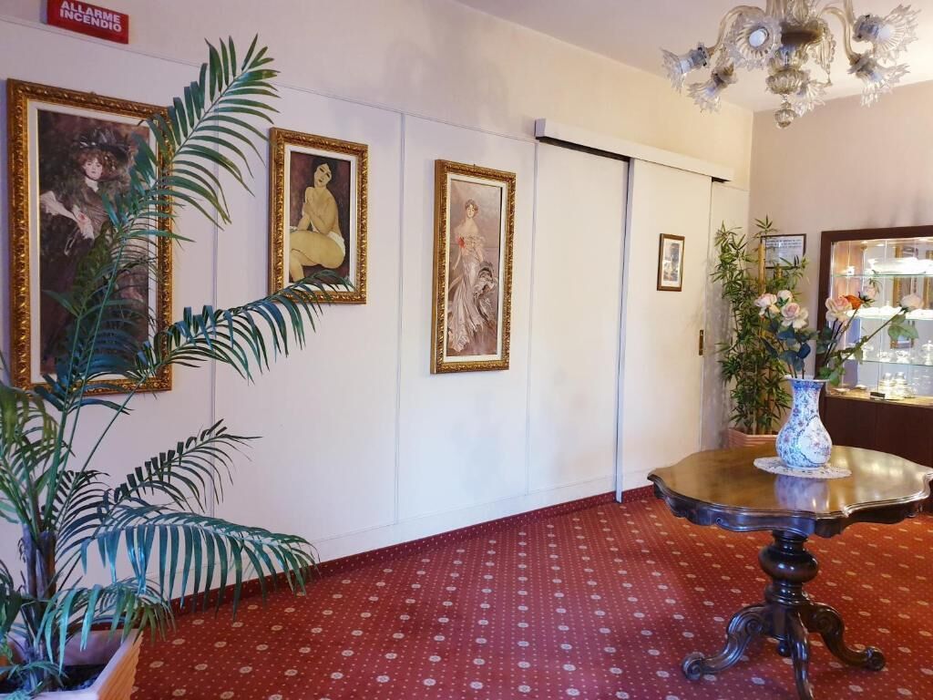 A hotel lobby with a red patterned carpet, framed artwork on the walls, a potted plant, and a table with a vase.