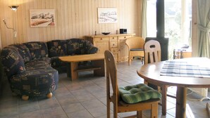 Una sala de estar acogedora con techo de madera, un sofá, una mesa de comedor y sillas. Hay dos cuadros en la pared y una ventana con cortinas.