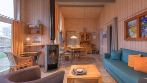 A cozy cabin interior with a wood-burning stove, a dining area, a sofa, and a large window. - Ferienhaus im Ferienpark Mueritz (Rechlin)