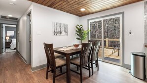 Un coin repas avec une table en bois, quatre chaises et une plante en pot. Une porte-fenêtre coulissante donne accès à un balcon d’où l’on peut admirer la vue sur les arbres.