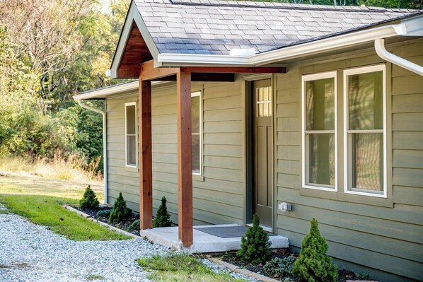 A single-story house with a covered porch, a front door, and a small front yard with a gravel path and young trees.
