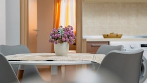 A modern dining area with a table set for two, a vase of flowers, and a kitchen in the background. - Levanta Cozy Apartment 3 in Kalavarda Rhodes (Rhodes)
