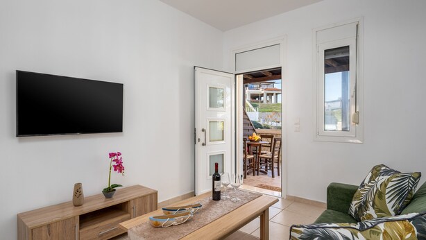 A modern living room with a flat-screen TV, wooden entertainment unit, a dining area with a table and chairs, and a green sofa. - Levanta Cozy Apartment 3 in Kalavarda Rhodes (Rhodes)