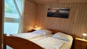 A wooden cabin bedroom with a bed, two bedside tables with lamps, a window with curtains, and a picture on the wall. - Ferienhaus im Skandinavischen Stil (Rechlin)