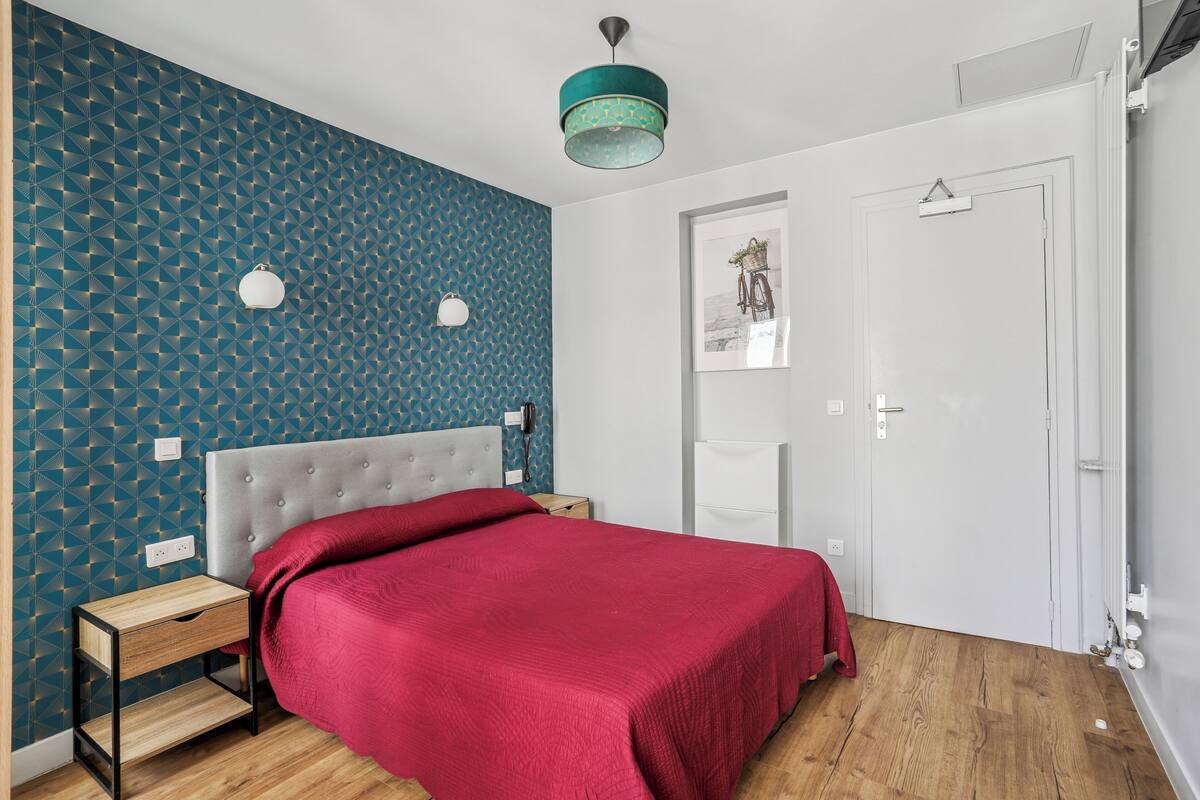 A bedroom with a patterned blue wall, a grey upholstered headboard, a red bedspread, a bedside table, a nightstand, a white door, and a framed picture on the wall.