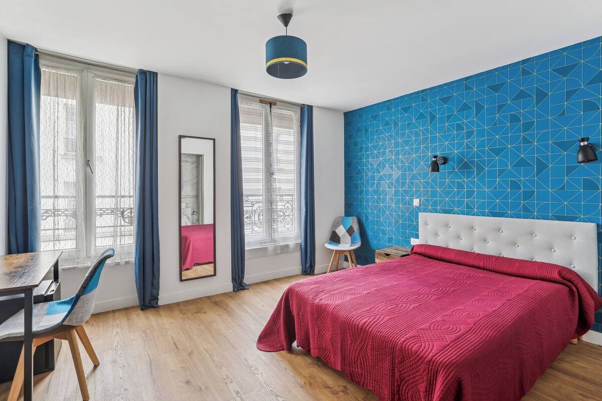 A bedroom with a large bed, a desk, and a chair. There are two windows with curtains, a mirror, and a blue geometric wall.