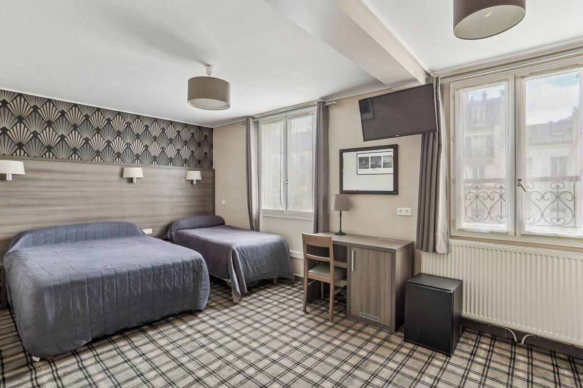 A hotel room with two beds, a desk, a chair, a radiator, and a window with a view of buildings.