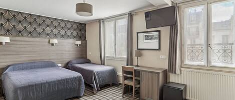 Quarto de hotel com duas camas, uma escrivaninha, uma cadeira, um radiador e uma janela com vista para edifícios.