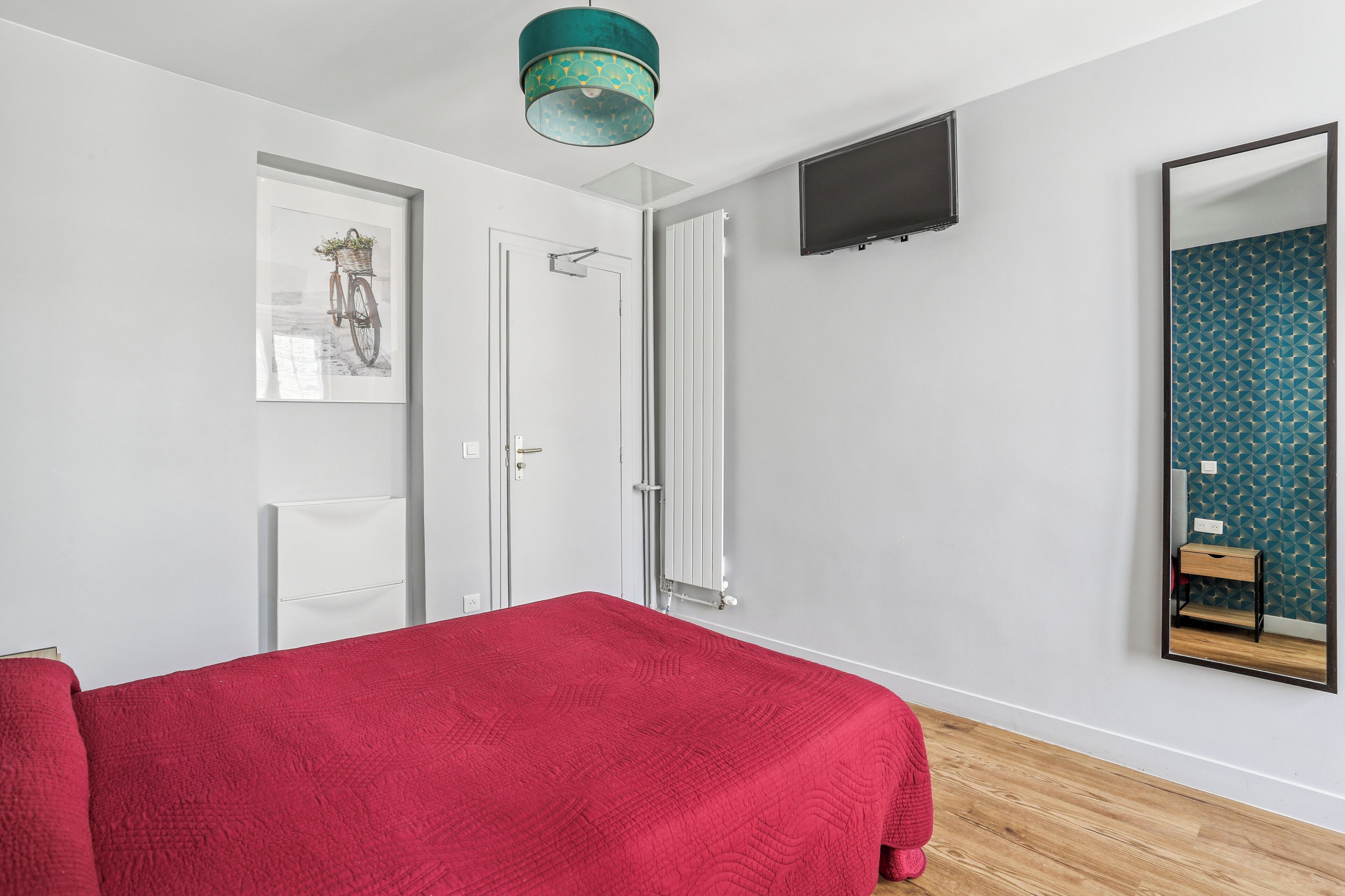 A bedroom with a red bedspread, a mounted TV, a mirror, and a door with a white frame.