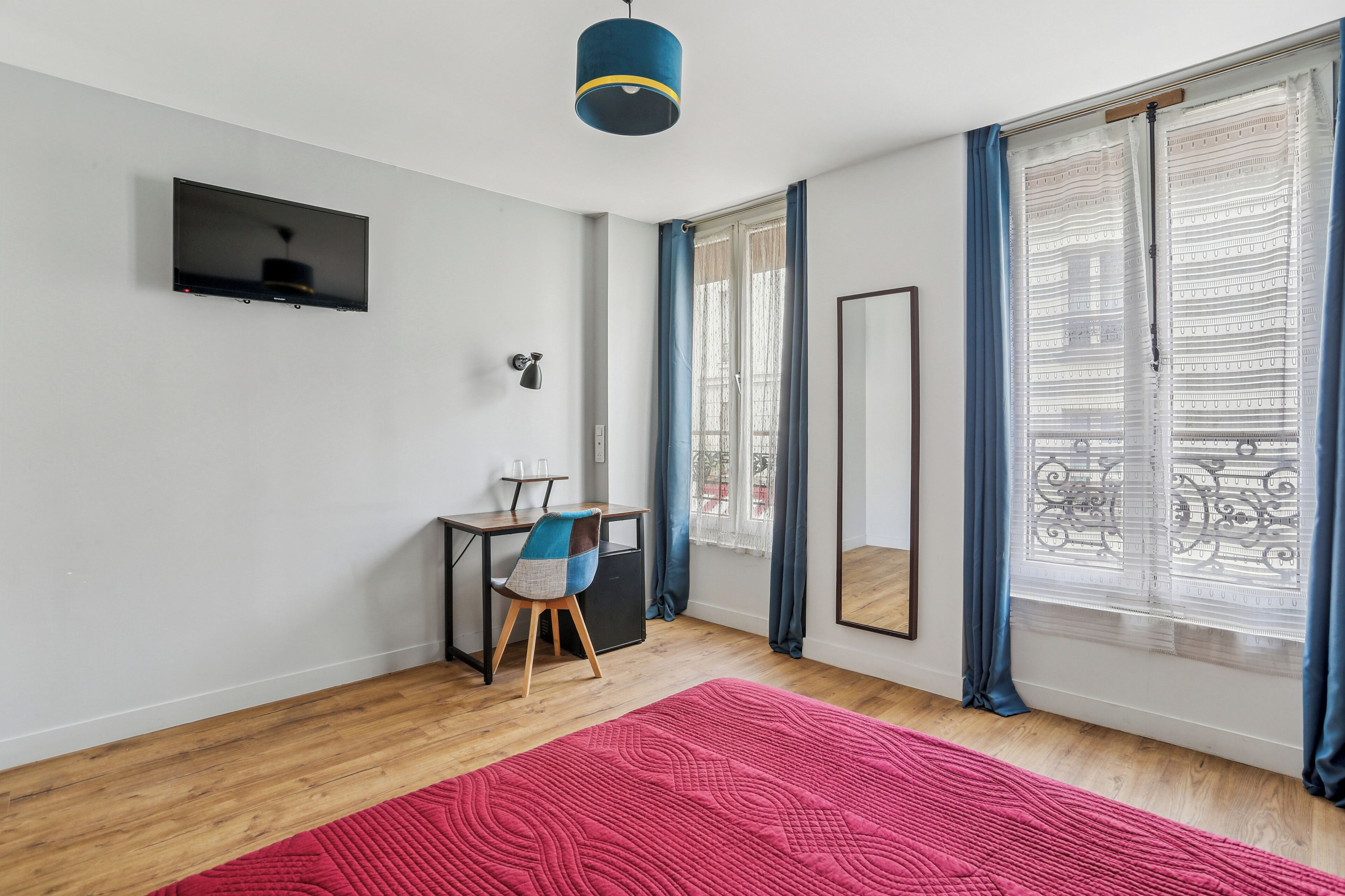 Una habitación moderna con piso de madera, un tapete rojo, un escritorio con silla, un televisor montado en la pared y un ventanal con cortinas.