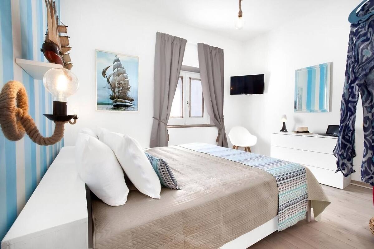 A bedroom with a bed, a desk, a chair, a ship painting, and a wall-mounted TV.