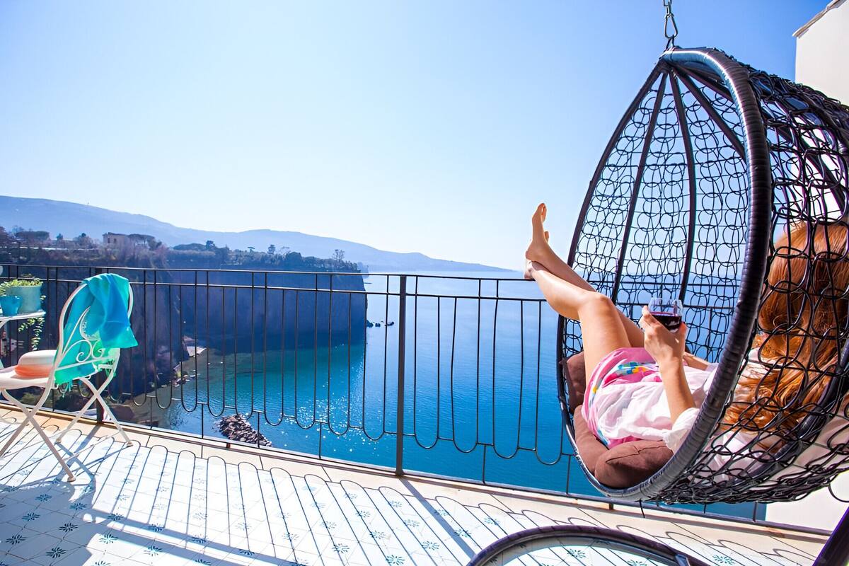 A person relaxing in a hanging chair by a balcony with a view of the sea and distant hills.