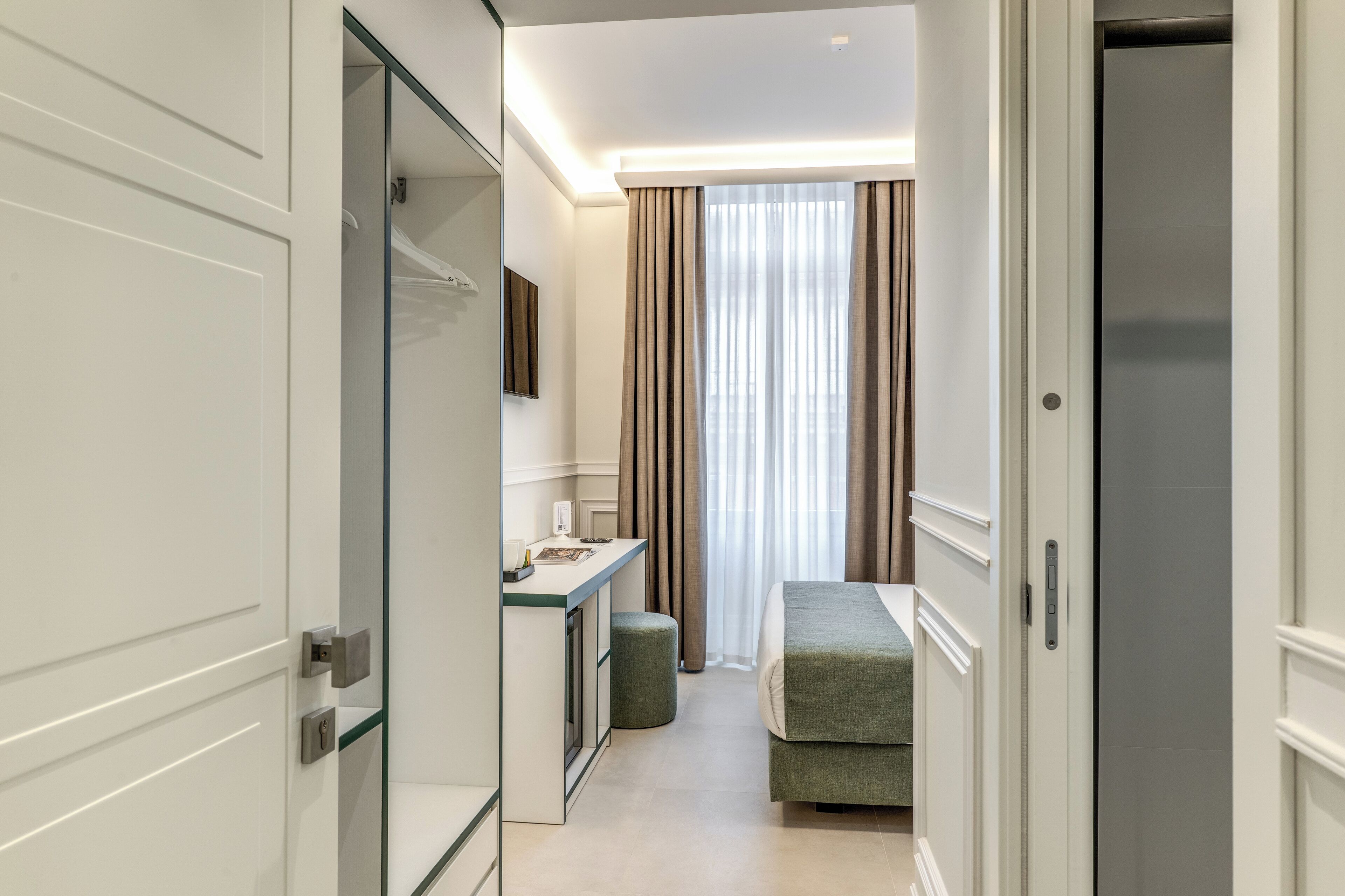 A hotel room with a bed, a small desk, a mirror, and a window with curtains.