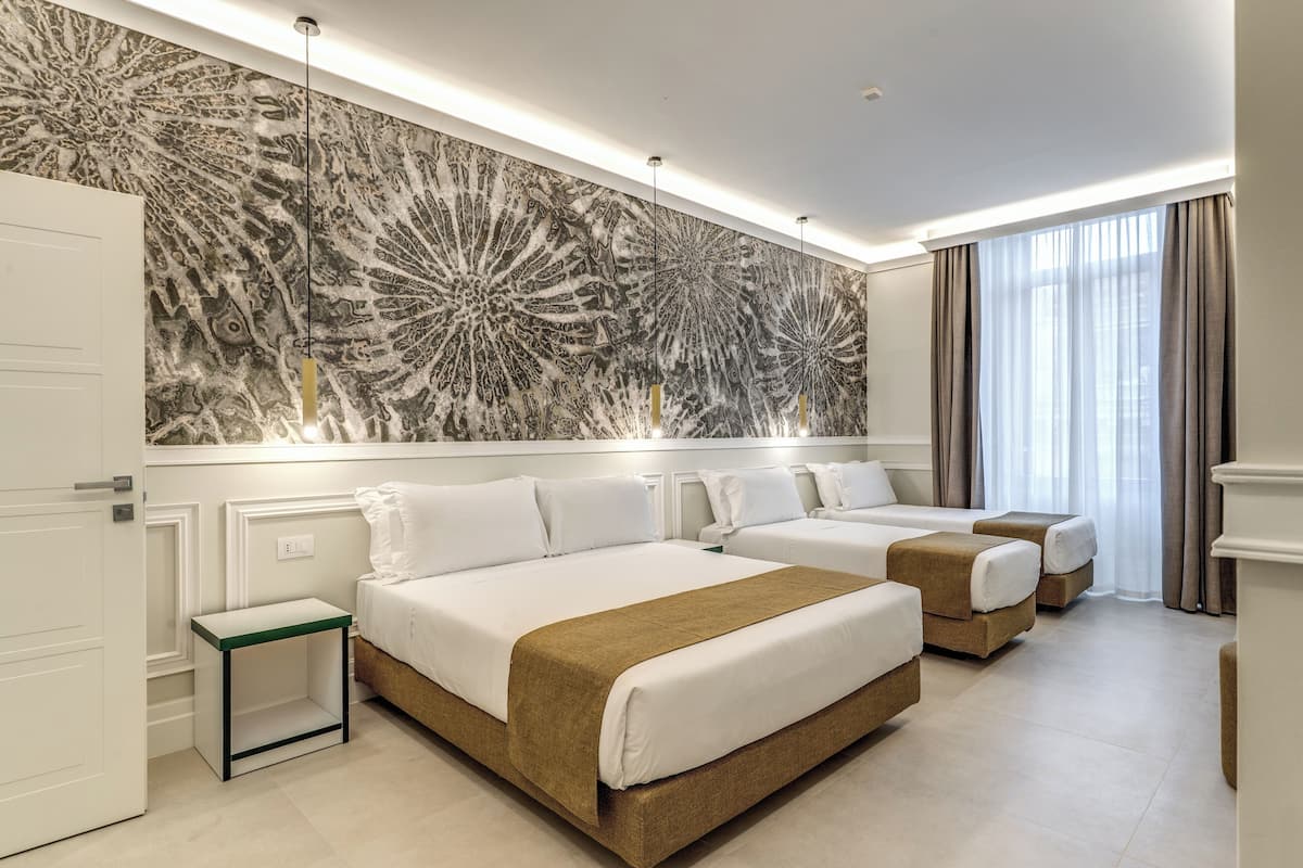 A hotel room with three beds, a large abstract wall mural, and a view of the city through sheer curtains.