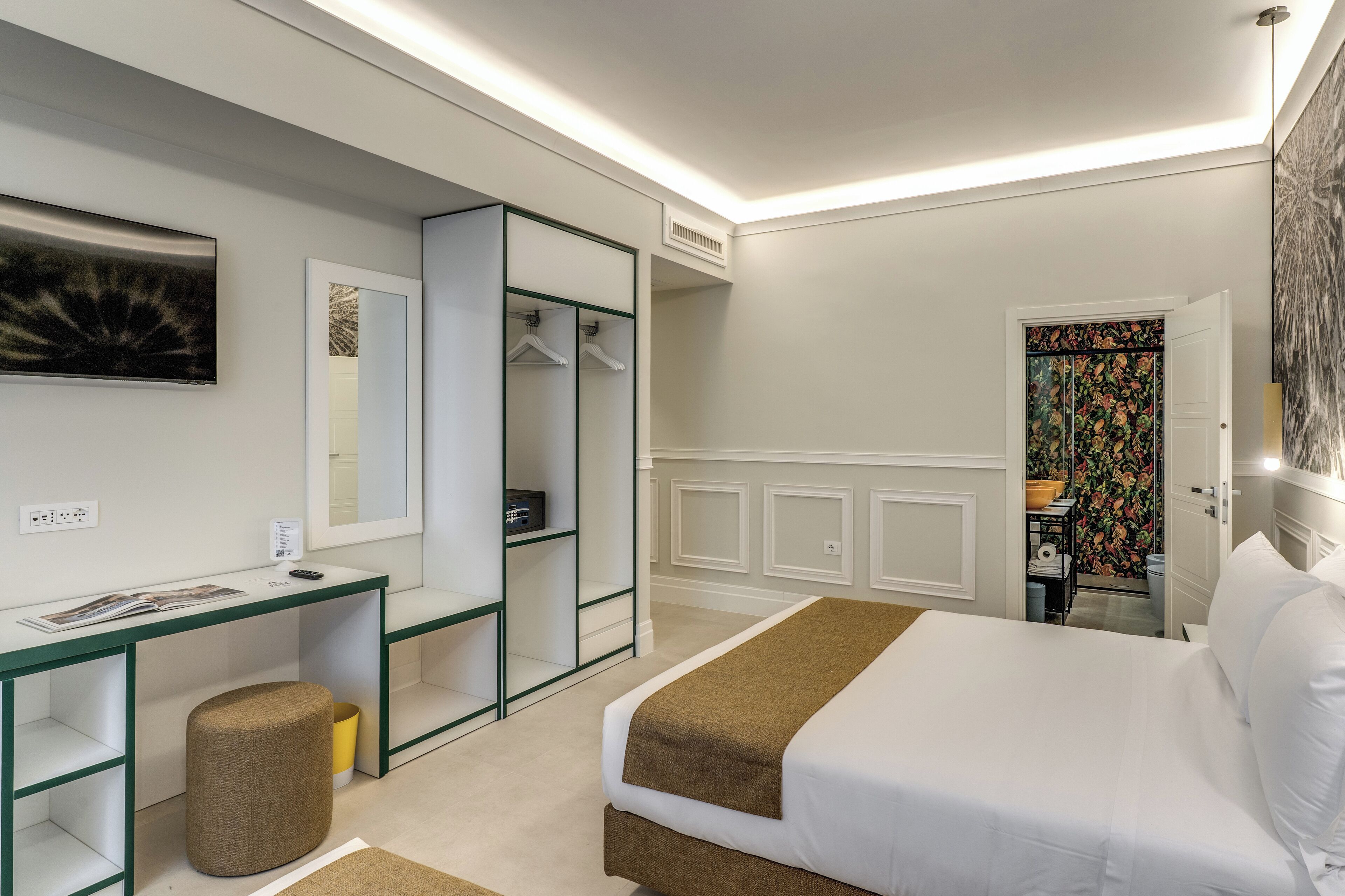 A modern hotel room with a bed, a desk, a wardrobe, and a bathroom visible through an open door.