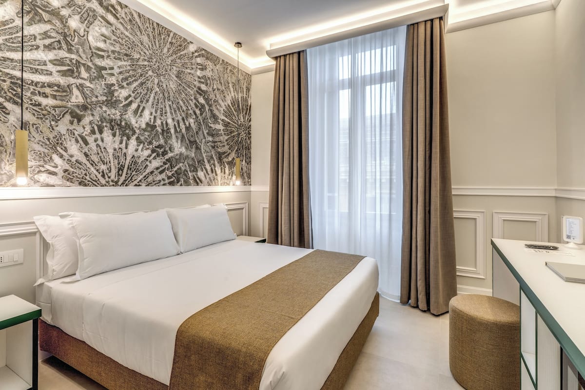 A hotel room with a large bed, a desk, a chair, a large abstract wall art, and a window with curtains.