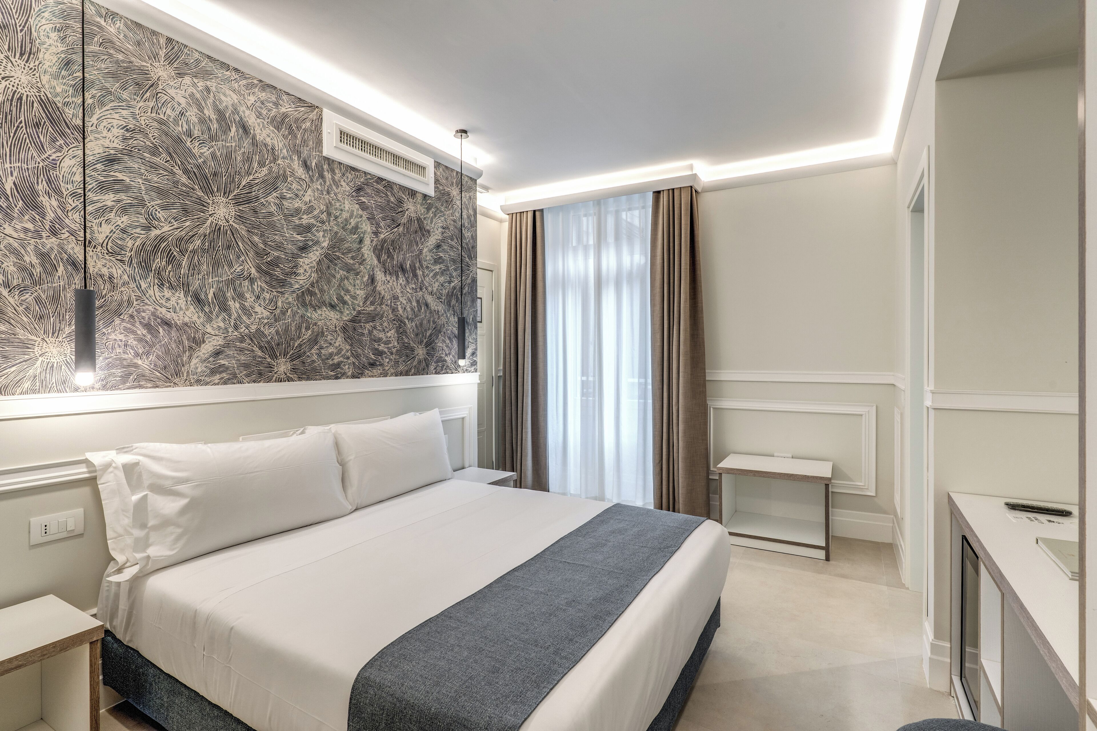 Quarto de hotel moderno com cama grande, mesa de cabeceira, escrivaninha e uma janela ampla com cortinas.