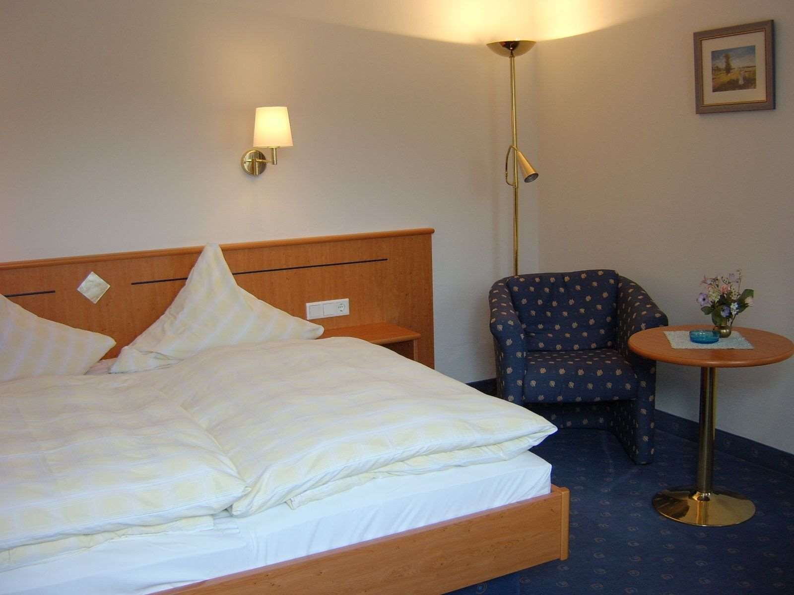 Quarto de hotel com uma cama, uma cadeira, uma mesinha com um vaso e um abajur.