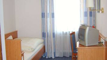 Una habitación de hotel pequeña con una cama individual, un escritorio con televisor, una silla y una ventana con cortinas.