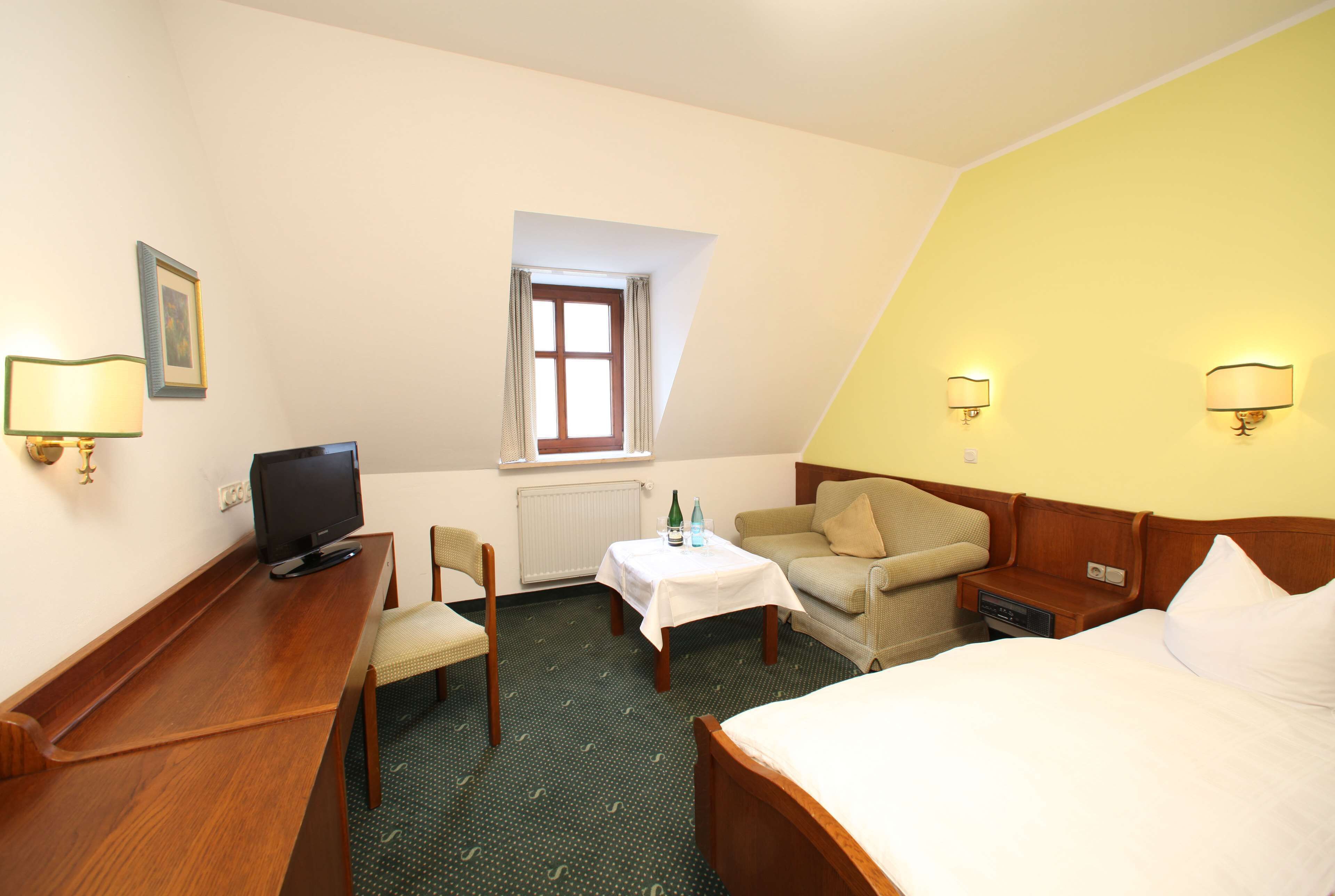 Une chambre d’hôtel avec un lit, un bureau, une chaise, une petite table avec une bouteille et des verres, un téléviseur et une fenêtre avec des rideaux.