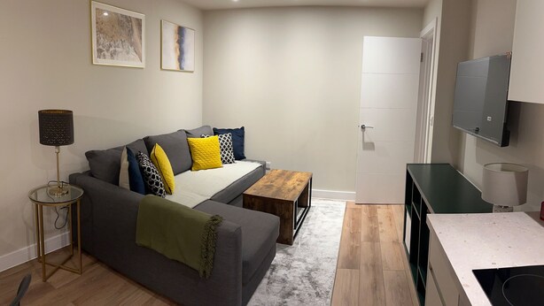 A modern living room with a grey sofa, a wooden coffee table, a small side table with a lamp, a TV mounted on the wall, and a door leading to another room. - Renovatedapartmentsleeps4-freeparking-tv (Brentford)