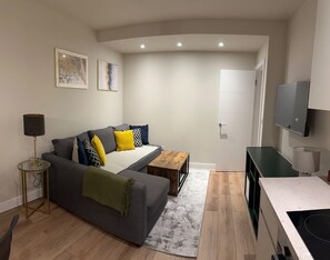A modern living room with a grey sofa, a wooden coffee table, a small side table with a lamp, a TV mounted on the wall, and a door leading to another room. - Renovatedapartmentsleeps4-freeparking-tv (Brentford)