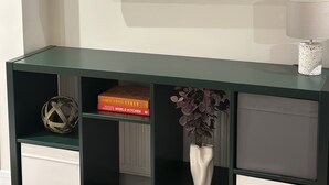 A modern living room with a mounted flat-screen TV, a dark green console table with storage drawers, and decorative items including a woven sphere and a vase with purple flowers. - Renovatedapartmentsleeps4-freeparking-tv (Brentford)