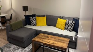 A modern living room with a grey sectional sofa, a wooden coffee table, and a grey rug. There are two abstract paintings on the wall and a small table with a lamp. - Renovatedapartmentsleeps4-freeparking-tv (Brentford)