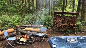 An outdoor hot tub surrounded by a fire pit and two chairs, set in a forested area with a stack of firewood nearby.