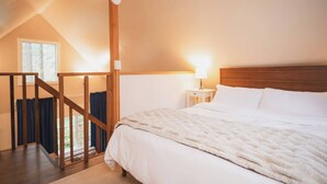 A bedroom with a large bed, a nightstand with a lamp, a wooden staircase, and a window with curtains.