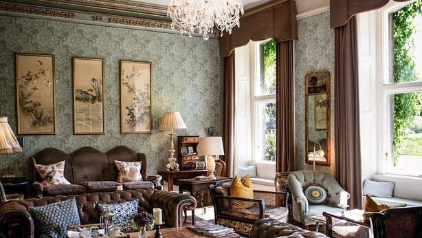 Ein traditionelles Wohnzimmer mit einem großen braunen Ledersofa, einem Holztisch und einer Kandelaberleuchte. An den Wänden hängen Bilder in Rahmen und ein Fenster mit Vorhängen.