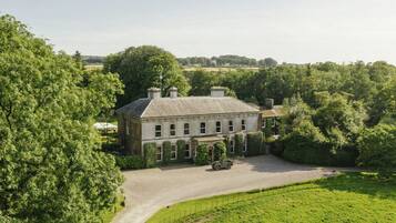 A large, historic building with a symmetrical facade, surrounded by lush greenery and a gravel driveway.