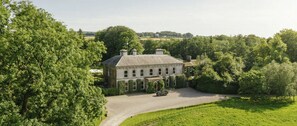 A large, historic building with a symmetrical facade, surrounded by lush greenery and a gravel driveway.