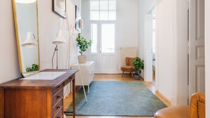 Un couloir avec une commode en bois, un miroir, deux lampes, un tapis vert, une chaise et une porte donnant sur une autre pièce.