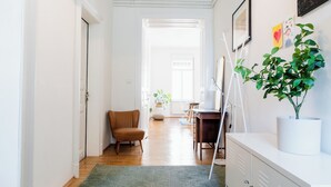 Un couloir avec un tapis à motifs géométriques, une suspension turquoise, un fauteuil marron, un meuble blanc orné d’une plante et une porte blanche.
