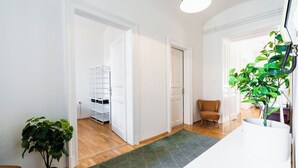 Une pièce spacieuse avec un plancher en bois, des murs blancs et un tapis à motifs verts. On y trouve une plante en pot, une chaise et un meuble blanc.