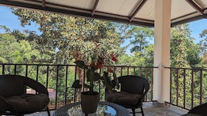 A covered outdoor seating area with wicker chairs and a table, surrounded by a metal railing, overlooking a lush green landscape. - Villa #248 (Somvarpet)
