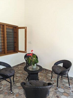 Un petit espace extérieur aménagé avec des meubles en osier et une table en verre, un pot de fleurs et une fenêtre dotée de volets en bois.
