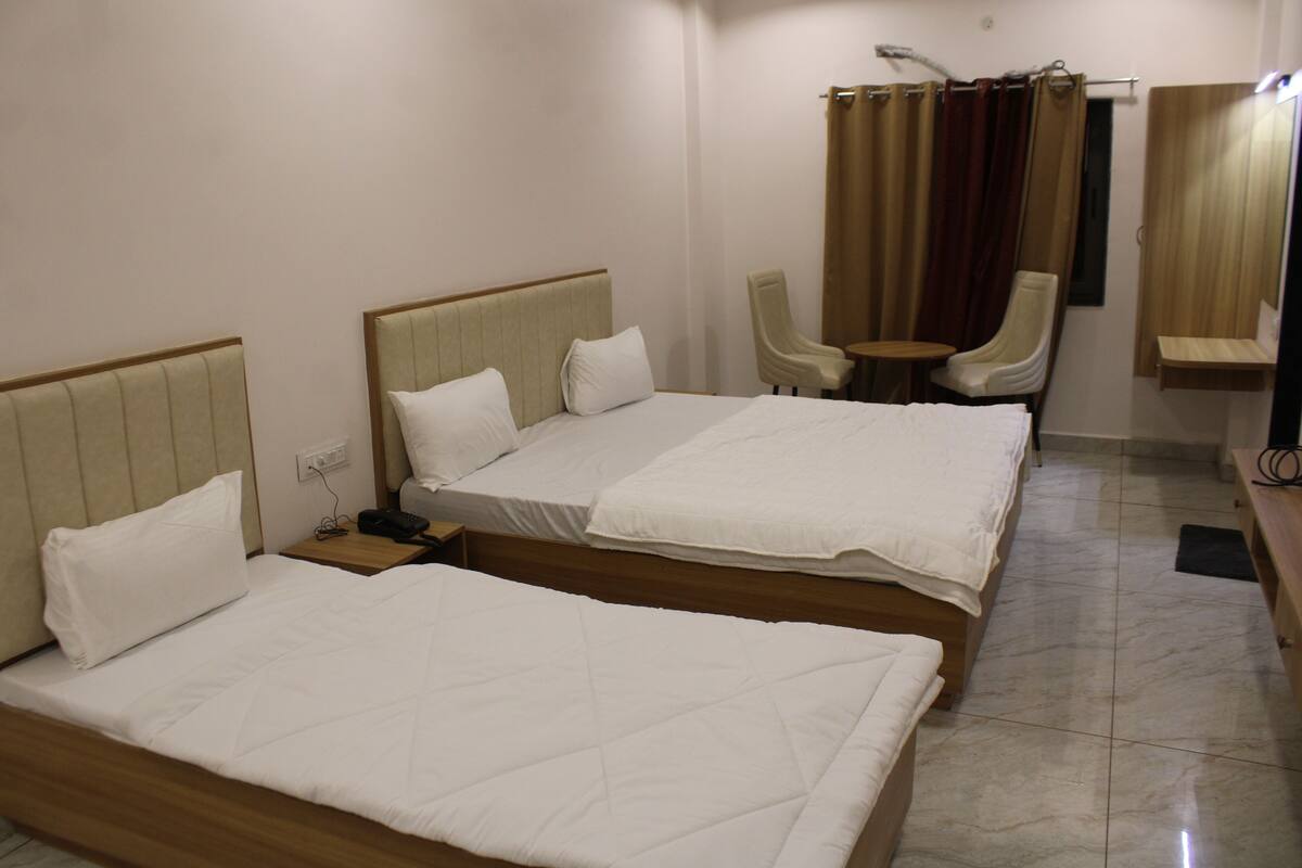 A hotel room with two beds, a small table, two chairs, a shower area, and a wooden bench.