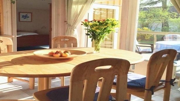 A wooden dining table with chairs, a vase of flowers, and a plate of fruit in a room with wooden walls and large windows. - Ferienhaus mit Sauna im Ferienpark Mirow (Mirow)