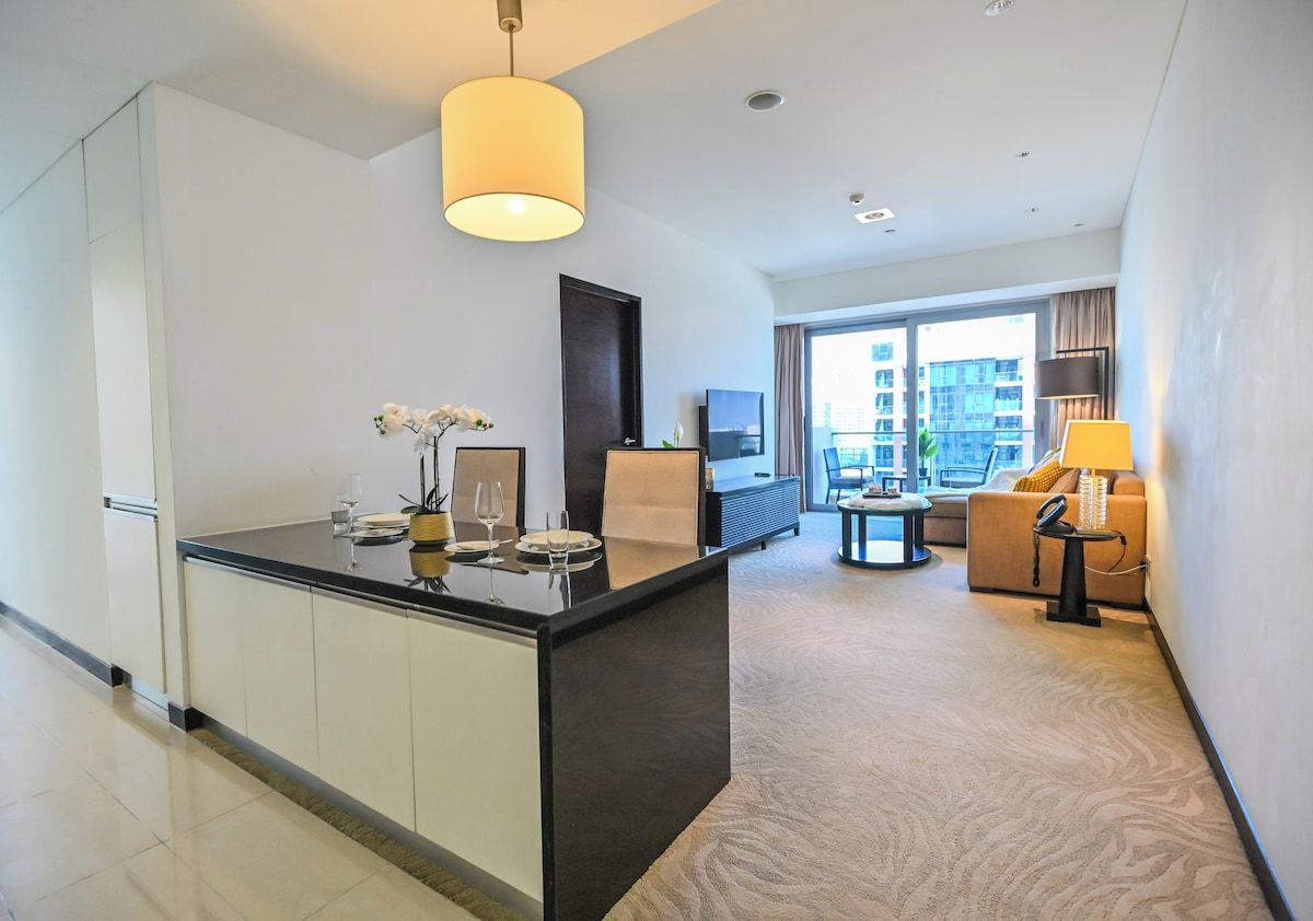 A modern hotel room with a kitchen area, a dining table, a sofa, and a large window offering a view of the city.